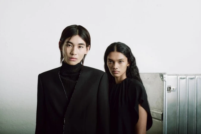 Two fashion models in black clothing pose in front of a textured white wall with a metallic case. Moody lighting and a studio setting.