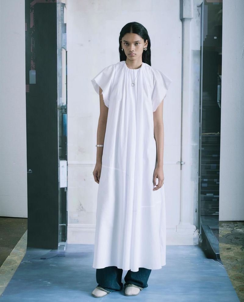 Model Nayonikaa Shetty wears a long white pleated dress over wide-leg denim jeans for MM6 Maison Margiela Holiday 2023.