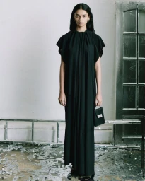 Model wearing a long black pleated dress holding a black handbag in an interior setting for MM6 Maison Margiela.