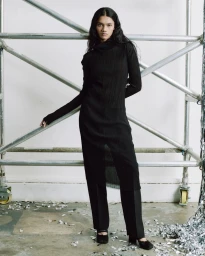 Model in long black dress and trousers leaning against industrial metal scaffolding in a studio setting