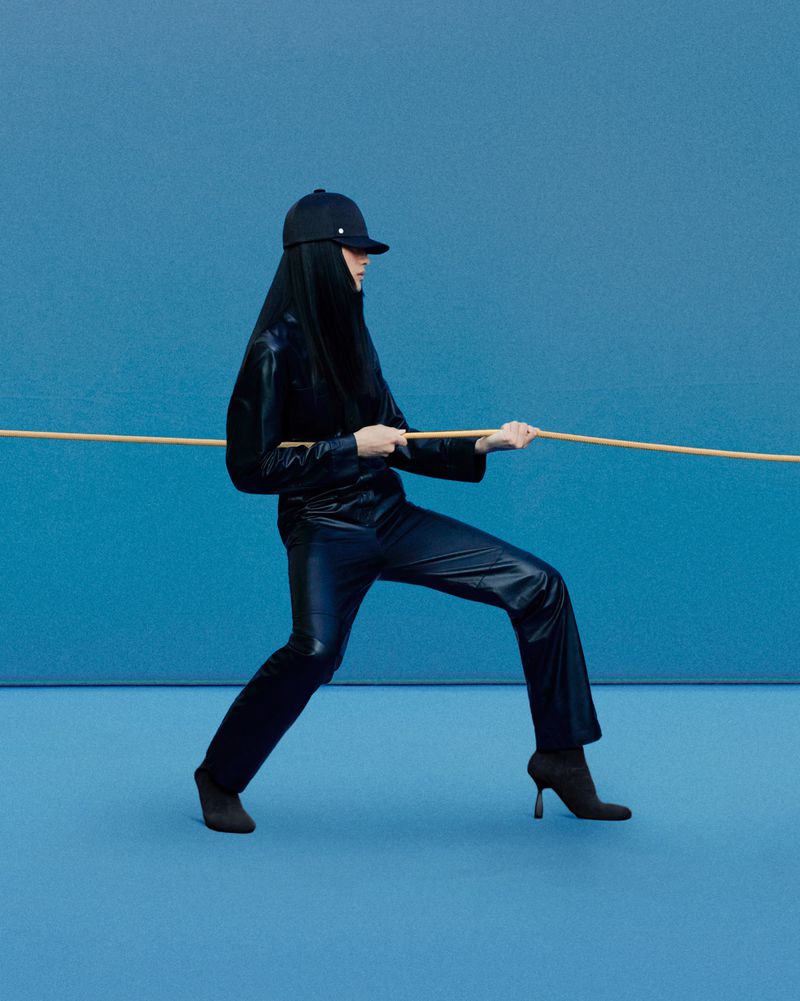 Woman in a black outfit pulling a rope against a blue background in a studio. The model is wearing a baseball cap and boots.