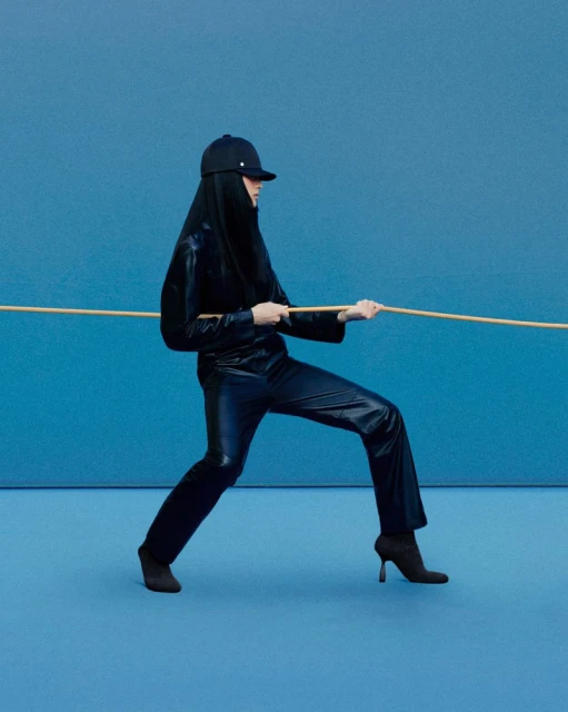 Woman in a black outfit pulling a rope against a blue background in a studio. The model is wearing a baseball cap and boots.