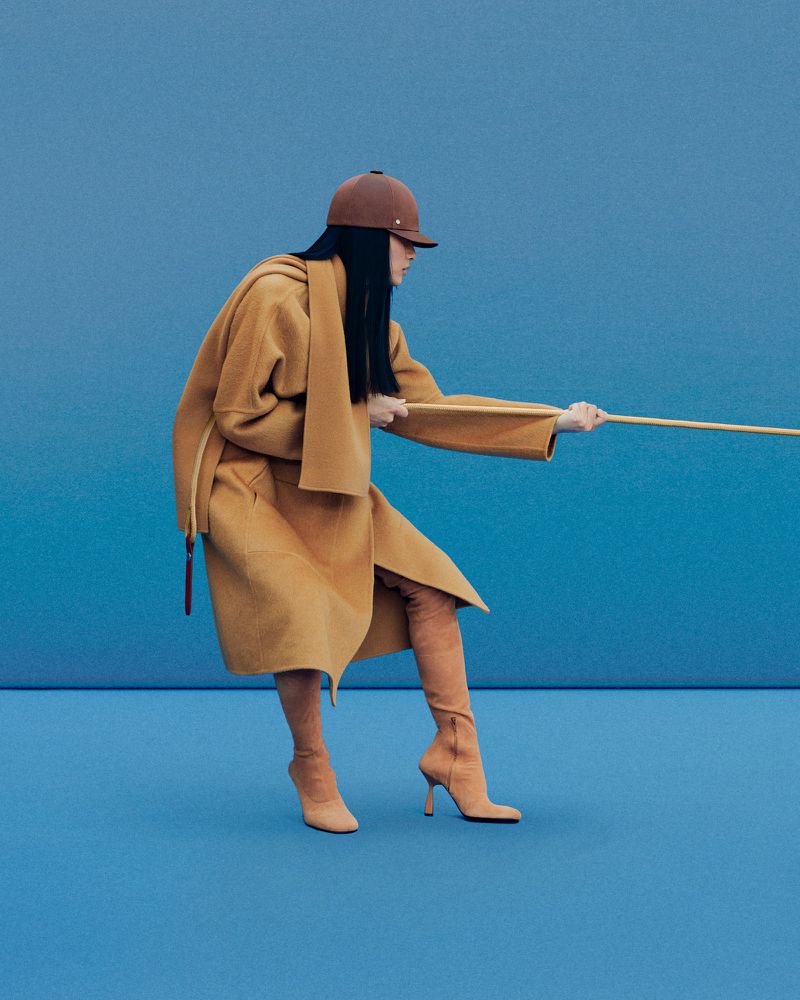 A woman poses in a tan coat, matching boots, and a riding hat against a blue backdrop, pulling a wooden pole in a fashion editorial.