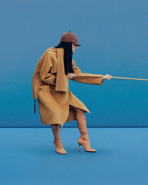 A woman poses in a tan coat, matching boots, and a riding hat against a blue backdrop, pulling a wooden pole in a fashion editorial.