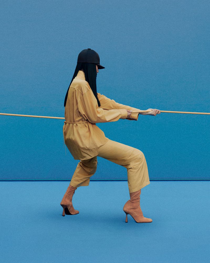 Woman in tan outfit and sock boots pulls rope against blue background. Fashion shot featuring single model in simple studio setup.