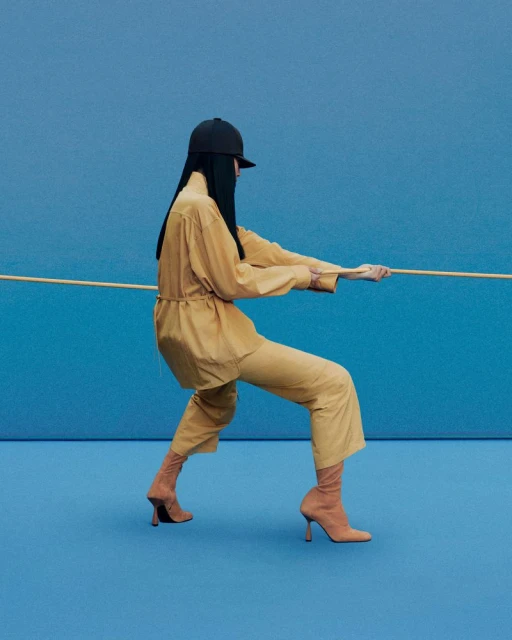 Woman in tan outfit and sock boots pulls rope against blue background. Fashion shot featuring single model in simple studio setup.