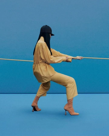 Woman in tan outfit and sock boots pulls rope against blue background. Fashion shot featuring single model in simple studio setup.