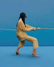 Model in yellow outfit wearing black cap and heeled boots pulling a rope against a blue studio background for Hermes.