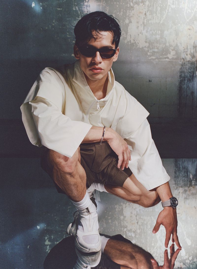 Young man in sunglasses crouches in a studio with white top, brown shorts, white sneakers, and gray watch, reflected on a shiny surface.