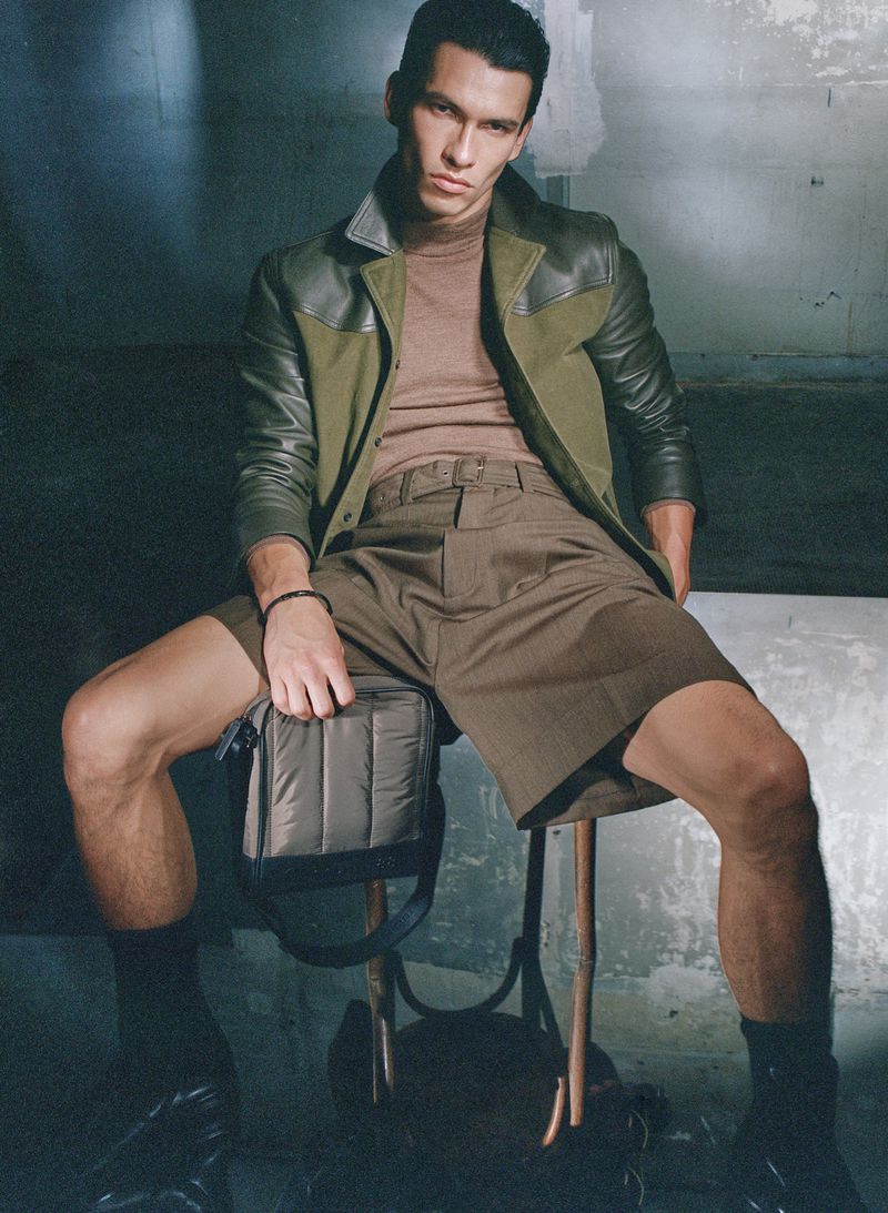 Fashionable man in shorts and jacket seated indoors, showcasing contemporary style and accessories in a posed studio shot.