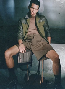 Male model in an olive green leather-panel jacket and tailored belted shorts seated on a stool holding a padded bag.