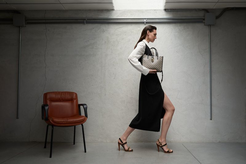 Model Angelika Baranska walking in a black slit dress and white shirt holding a patterned tote bag next to a brown leather chair.