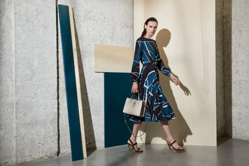 A slender woman walks in a fashion shot wearing a patterned blue dress with a handbag, with a geometric modern backdrop.