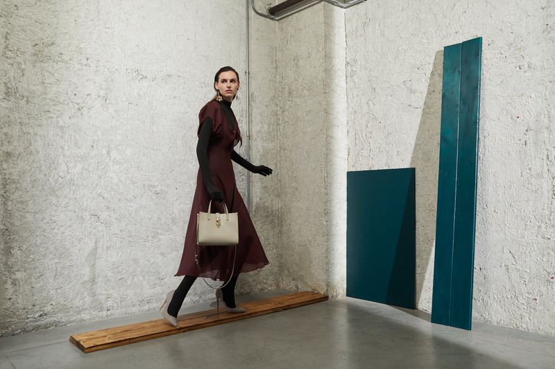 Fashion editorial shot: a stylish adult female model in a maroon dress walks with a handbag in a minimalist studio setting.