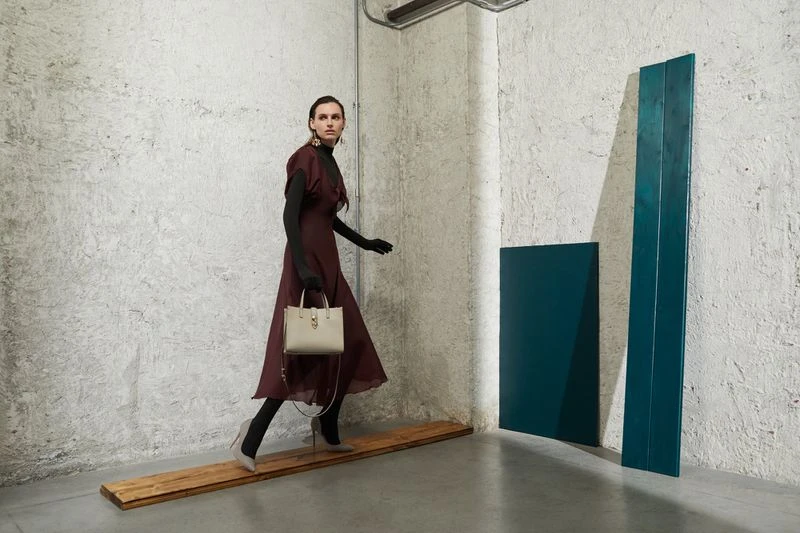 Fashion editorial shot: a stylish adult female model in a maroon dress walks with a handbag in a minimalist studio setting.