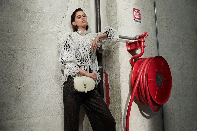 A woman in a white lacy top and dark pants poses against a concrete wall, with a red fire hose reel visible in the background.
