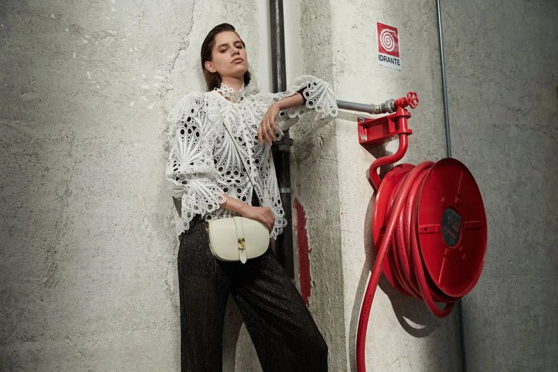 A woman in a white lacy top and dark pants poses against a concrete wall, with a red fire hose reel visible in the background.