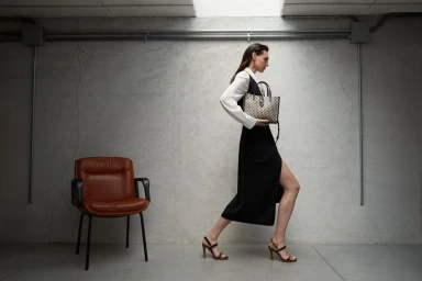 Model Angelika Baranska walking in a black slit dress and white shirt holding a patterned tote bag next to a brown leather chair.