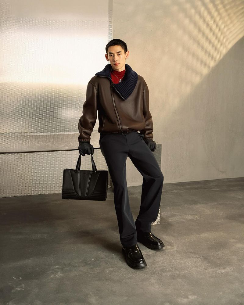 Fashionable Asian man in a brown leather jacket and black pants poses with a black tote bag in an indoor setting with sunlight.