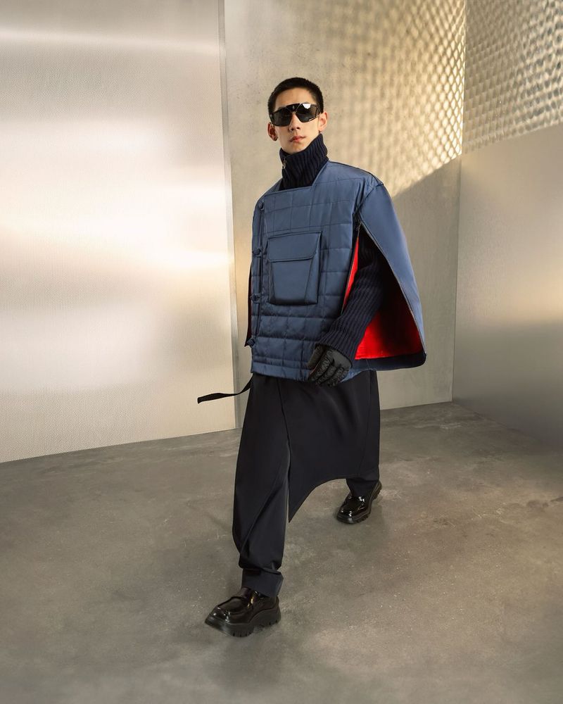 An adult male fashion model strides confidently in a photo studio, wearing a blue quilted vest, dark turtleneck, draped pants, and sunglasses.