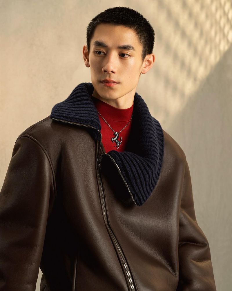Portrait of a young Asian man wearing a brown leather jacket and a silver pendant with a horse logo on a neutral background.