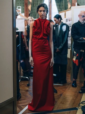 A fashion model in a red gown backstage at a fashion show, surrounded by crew members in a backstage setting.