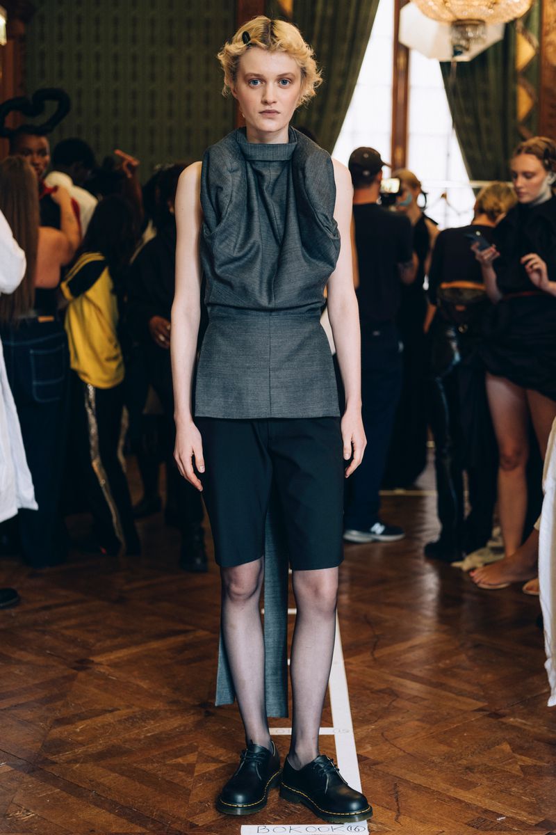 Fashion runway model in gray sleeveless top and black shorts poses with confidence in a brightly lit indoor space during a show.