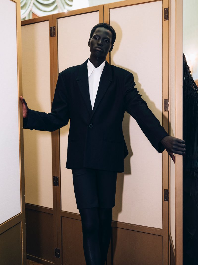 Fashionable Black model strikes a pose between room dividers, sporting a black suit and white shirt in an indoor setting.