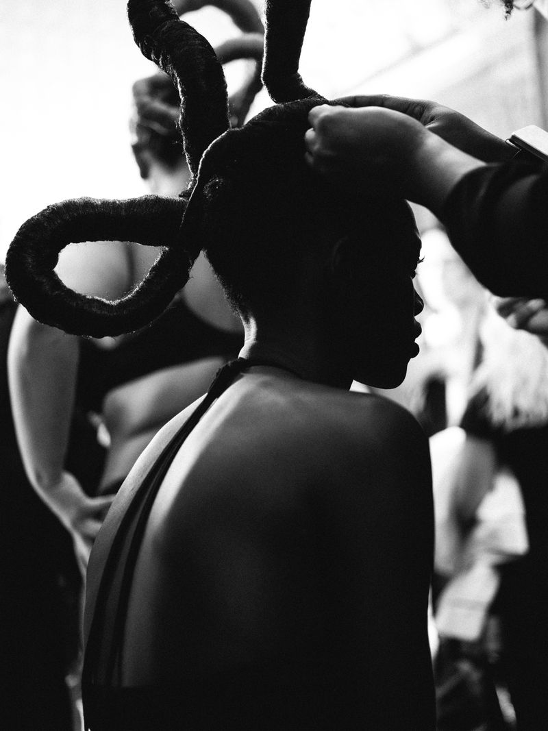Black and white image of a dark-skinned adult female having her hair styled into horn-like shapes, assisted by a person's hands.