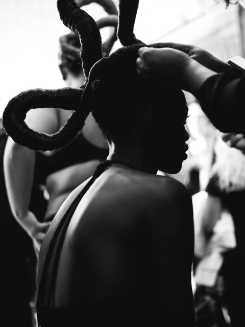 Black and white image of a dark-skinned adult female having her hair styled into horn-like shapes, assisted by a person's hands.