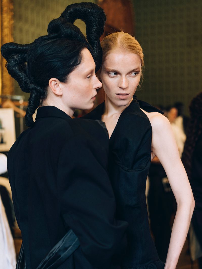 Two fashion models in black dresses, one with extravagant hair. Backstage shot of women with editorial hair and makeup.