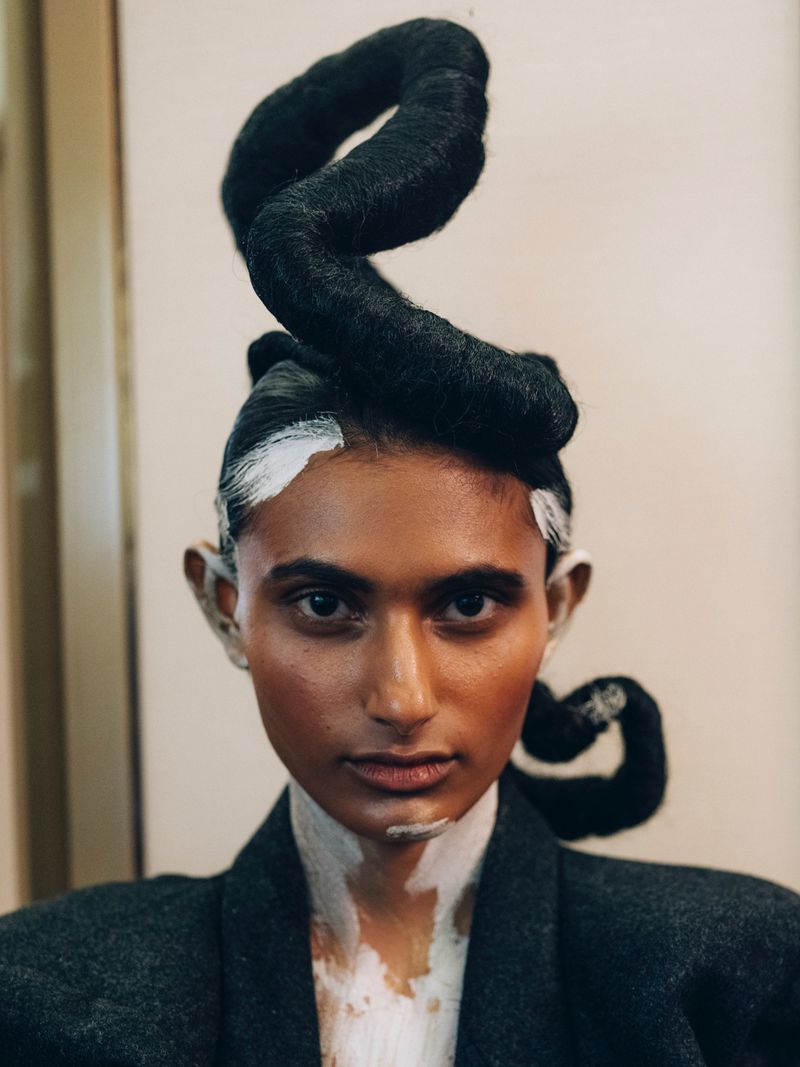 Close-up studio shot of a person with dark skin, creative hairstyle, white makeup, wearing a grey suit jacket, portrait orientation.