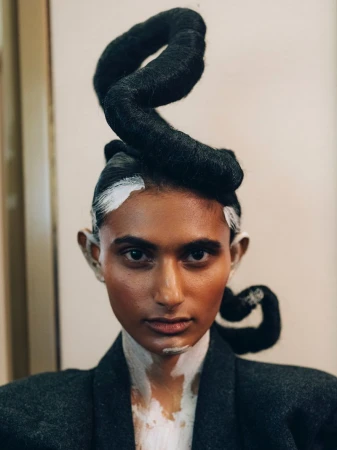 Close-up studio shot of a person with dark skin, creative hairstyle, white makeup, wearing a grey suit jacket, portrait orientation.