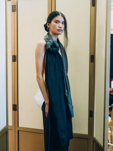 Portrait of an adult female with dark hair, wearing a dark sleeveless dress with gray shawl, posing in an indoor setting with light walls.