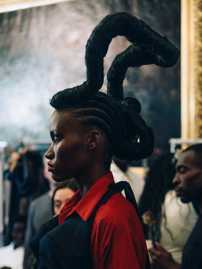 Striking side-profile portrait of a woman with elaborate braided updo wearing a red-collared shirt in a backstage fashion show setting.