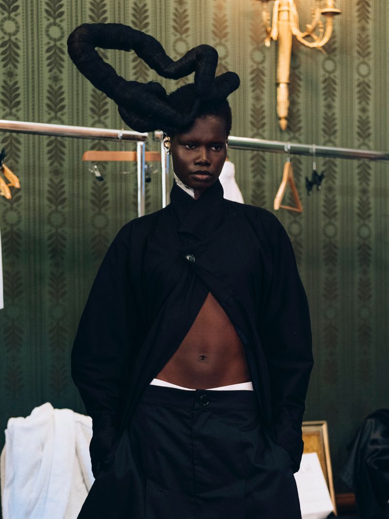 Woman in draped black coat and sculptural updo against patterned wallpaper and clothes rack, fashion editorial, indoor studio setting.