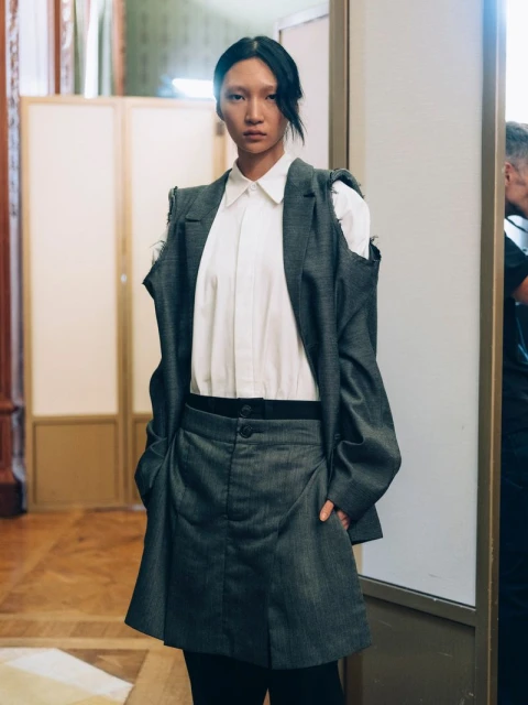 Attractive woman in a blazer and skirt, hands in pockets, stands in front of a white wall in an indoor environment with neutral light.