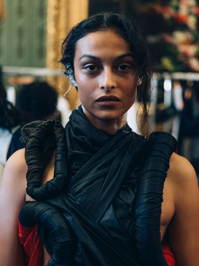 Close-up portrait of a model with dark hair wearing a draped black top in front of a floral-wallpapered dressing room backstage.