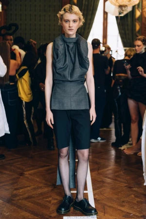 Model in a grey sleeveless high-neck top and black shorts backstage for TORISHÉJU S/S 24 in Purple Magazine