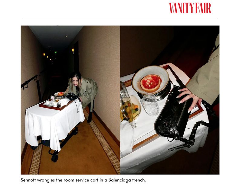 A woman in a trench coat pushes a room service cart down a hotel hallway. The cart features a tray with various food items.