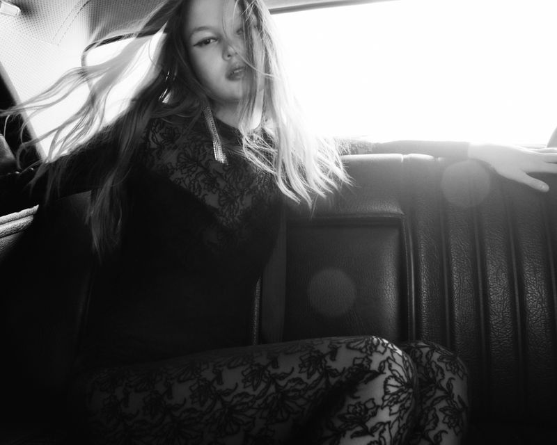 Young woman with long blond hair wearing a black floral lace dress inside the backseat of a car with bright window light.