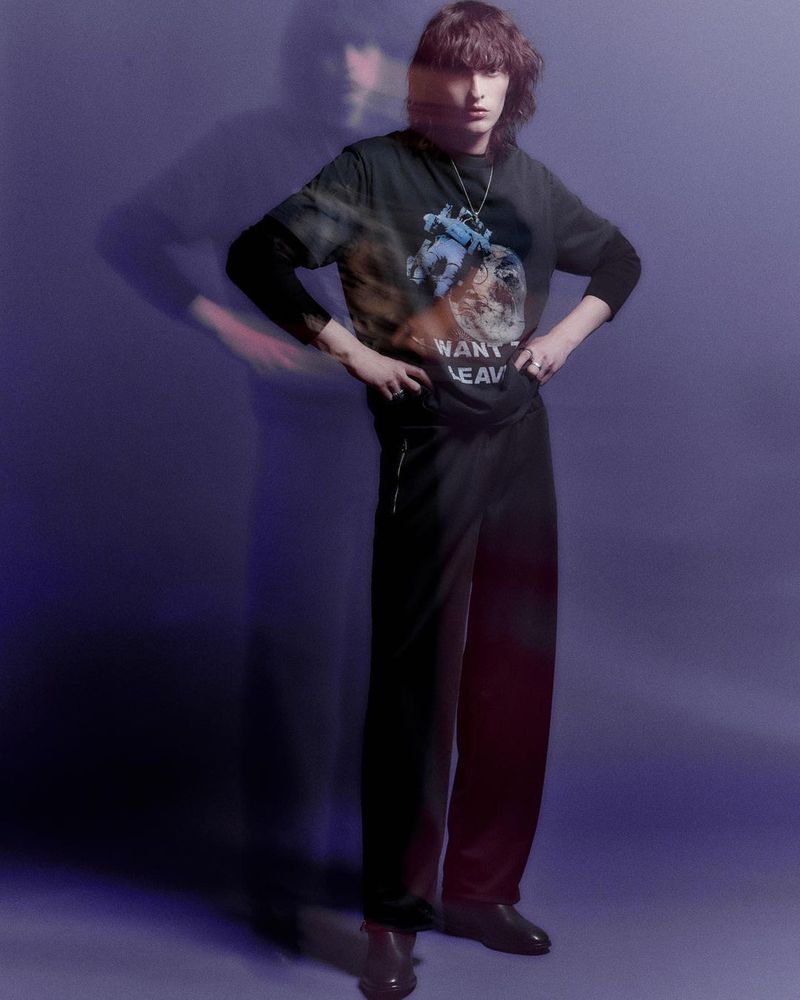 Full-length portrait of a young adult wearing a black t-shirt with graphic print, black pants, and brown shoes, with blurred motion effect.