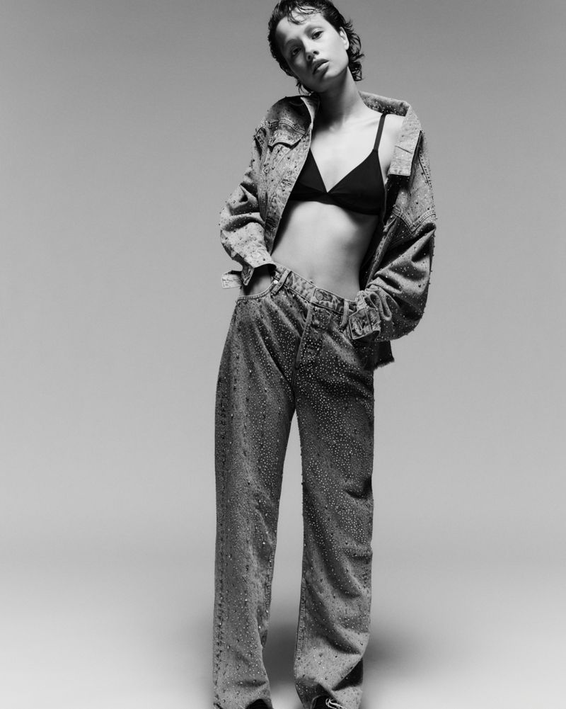 Monochrome full shot of a young adult woman in a denim jacket, bra, and jeans, posing in a photo studio against a light background.