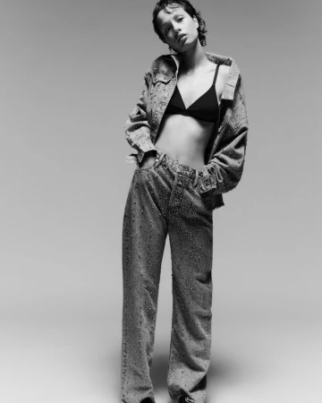 Monochrome full shot of a young adult woman in a denim jacket, bra, and jeans, posing in a photo studio against a light background.