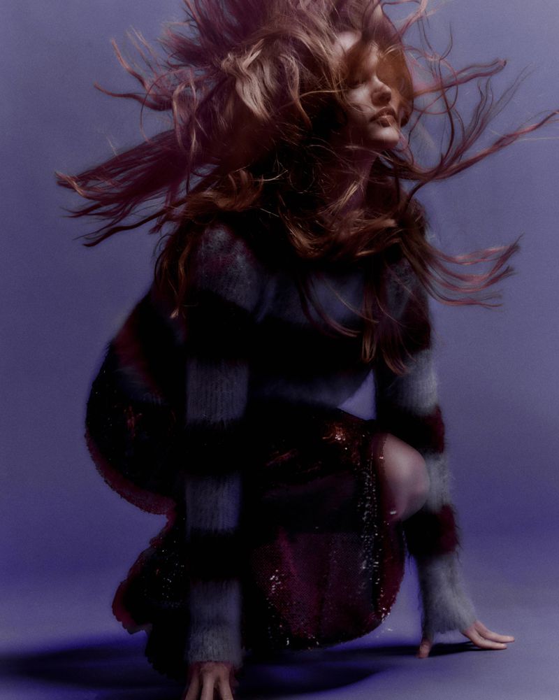 Dynamic fashion portrait of a woman with long, flowing hair in a striped top and shimmering skirt, crouching against a blurred purple backdrop.