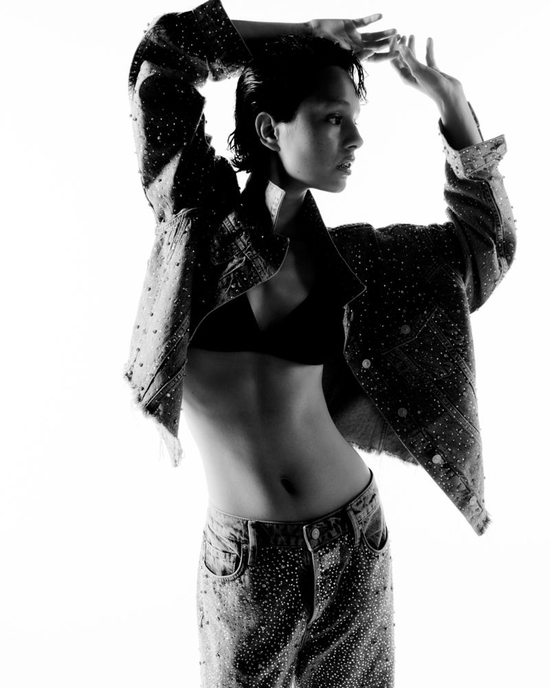 Black and white fashion portrait of a young woman with short hair wearing a denim jacket and jeans with embellishments, posing in a bright, stark studio.