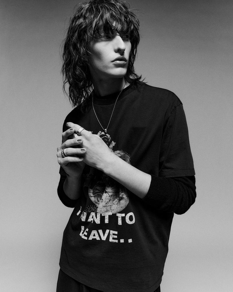 Monochrome portrait of a young adult male with long hair wearing a graphic tee and jewelry, looking off-camera against a plain backdrop.