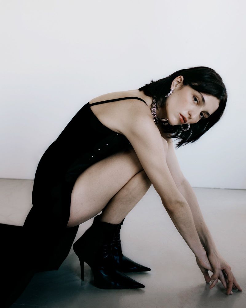 A slender young woman with dark hair poses indoors wearing a black dress, jewelry, and black stiletto boots against a white background.