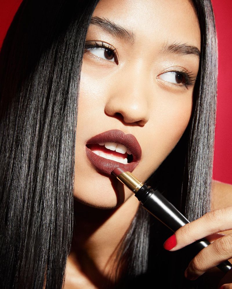 Close-up of an Asian woman applying dark red lipstick, showcasing beauty, makeup, and cosmetics against a vibrant red backdrop.