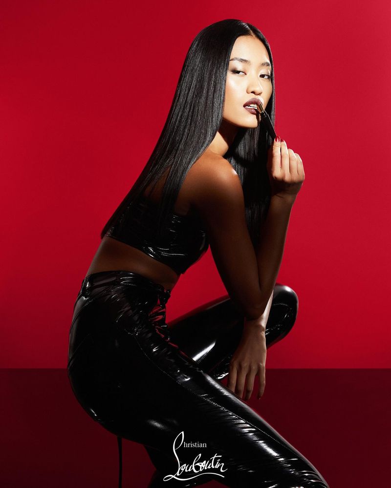 Portrait of a young Asian woman with glossy black hair, dressed in a black latex top and pants against a vibrant red backdrop.